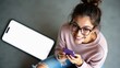 © Leonardo - Mujer joven sonriente con gafas en ropa informal sentada cerca de un teléfono inteligente gigante con pantalla blanca en blanco, usando el móvil