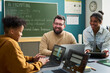 © pressmaster - Caucasian man smiling and interacting with Black teenage girl and Black teenage boy using laptops in classroom setting, chalkboard with AI prompting instructions visible in background