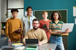 © pressmaster - Group of diverse teenagers standing with Caucasian man holding digital tablets in classroom, teenagers smiling and looking at camera, teacher sitting at desk with laptop, chalkboard in background