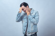 © Daniel - Asian man in denim jacket touching his cheek with painful expression, standing in studio against plain background, showing toothache, jaw pain, and dental problem