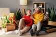 © sementsova321 - Two older adults sit together on the floor, sharing smiles amidst unpacked items, plants, and warm light from a lamp in their new living room