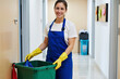 © Alex & M Studio - Portrait of a happy female janitor and professional cleaner in a uniform and gloves smiling while holding her equipment in an office hallway