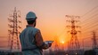 © EarthWalker - Electrical engineer technician in hard hat with a tablet computer device monitoring high voltage transmission tower power grid lines at sunset