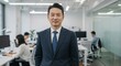 © MstShilpi12 - Confident asian businessman in a sharp suit stands in a modern well lit office environment with colleagues working in the background