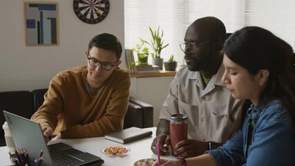 Three biracial IT specialists sitting at desk, looking at laptop screen discussing new app for smartphones