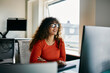 © Flamingo Images - Young Professional Woman Working Smiling in Modern Office Environment. She is wearing an orange sweater, beautiful curly hair and black glasses. She is looking intently at a screen.