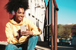 © halayalex - Beautiful woman with afro curls hairstyle.Smiling model in yellow hoodie.Sexy carefree female enjoying listening music in wireless headphones.Posing on street background at sunset.Holds phone