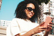 © halayalex - Beautiful woman with afro curls hairstyle.Smiling model in white hoodie. Sexy carefree female posing on the street background in sunglasses. Looking at smartphone screen, using apps
