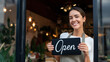 © dinatychynska - Smiling woman business owner holding open sign at storefront entrance. Happy female entrepreneur welcoming customers to new shop opening with warm interior lighting and plants.