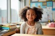 © id512 - Cute african american schoolgirl smiling at desk in colorful elementary classroom. Young female student with curly hair looking at camera during lesson indoors