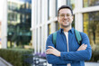 © Liubomir - Young man with glasses and backpack stands on a university campus, arms crossed, smiling confidently at the camera-casual, modern student portrait conveying success and positivity