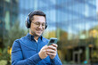 © Liubomir - Man with glasses and headphones smiling outdoors in an urban street, casually using his smartphone to stream music and chat, relaxed and confident in daylight city setting