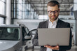 © anatoliycherkas - Modern bearded man in glasses and suit vehicle sales consultant using laptop inside car dealership