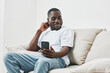 © SHOTPRIME STUDIO - Young Black man enjoying music on his smartphone while relaxing on a light colored couch in a modern living room, conveying a sense of happiness and comfort