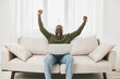 © SHOTPRIME STUDIO - Joyful man celebrating success while using a laptop in a bright living room setting, wearing a cozy sweater, showcasing happiness and accomplishment