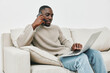 © SHOTPRIME STUDIO - Young Black man in cozy sweater engaging in phone conversation while working on laptop at home, showcasing a warm and productive atmosphere