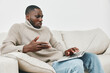 © SHOTPRIME STUDIO - A thoughtful African American man in a cozy beige sweater sitting on a couch, engaging with a laptop, embodying curiosity and contemplation in a minimalist living space