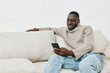 © SHOTPRIME STUDIO - Smiling young Black man in a cozy beige sweater engaging with his smartphone while sitting on a light colored sofa, showcasing modern relaxation and digital lifestyle