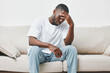 © SHOTPRIME STUDIO - A young African American man sitting on a couch, looking stressed and thoughtful, dressed in a plain white t shirt and blue jeans, reflecting deep emotions