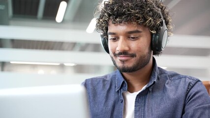 Confident businessman or IT specialist in wireless headphones works on a laptop sitting at workplace in business office. Focused coder developer is programming, busy with project on computer. Close up