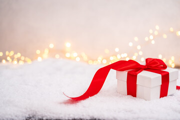Wall Mural - festive white gift box tied with a red ribbon, resting on snow with blurred golden lights in the bac