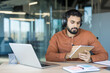 © Liubomir - Young man with headphones works on a laptop in a modern glass-walled office, focused on taking notes in a notebook while studying or attending an online course for professional growth