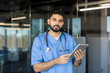 © Liubomir - Confident male healthcare professional in blue scrubs with stethoscope, holding a tablet in a modern clinic, looking at camera tech-savvy physician ready to consult and care