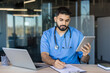 © Liubomir - Male doctor in blue scrubs and stethoscope reviews patient data on a tablet while taking notes on a clipboard at a clinic desk, focused and professional