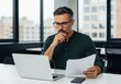 © Alif - Focused businessman analyzing documents on laptop in modern office setting contemplating financial strategy and business growth.