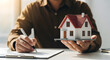 © Dewi - Business person holding a model house and writing on a notepad representing real estate investment or property planning in a professional setting