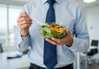 © Arrhman - A businessman in a shirt and tie holds a plastic bowl of fresh salad with a fork in an office setting.