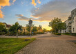 © tl6781 - quiet suburban street glows under a golden sunset sky, with soft clouds, trees, and modern houses lining the calm, cobblestone-paved road