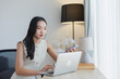 © Tj - Asian businesswoman working remotely from a home office, skillfully multitasking between her laptop and smartphone while sitting at a wooden desk near a window