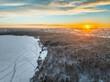 Snow-covered forest on lake shore with ice at sunset and the city on horizon, auerial view