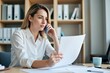 © WoodHunt - A white woman is speaking on the phone while reviewing documents at her desk in a bank office. The background shows shelves with files and a computer