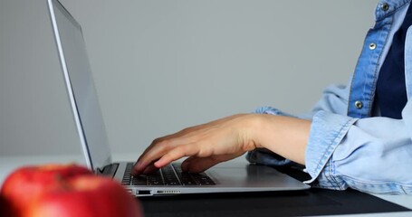 Woman programmer coding on laptop keyboard, wearing denim shirt, sitting at workspace with fresh apple nearby, focusing on software development project against minimalist gray background