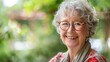 © Chandara - A smiling elderly woman with gray hair, wearing glasses, standing outdoors in a garden with greenery in the background.
