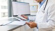 © Enigma - Male doctor wearing a white lab coat and stethoscope, carefully reviewing patient documents and healthcare paperwork while working at a desk in a medical office with a computer