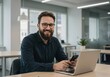 © sujon1638 - Smiling bearded man with glasses holding a smartphone while sitting at a desk with a laptop in a modern office environment