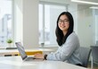 © sujon1638 - Young woman with glasses smiling while working on a laptop computer at a modern office desk with large windows