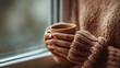 © Karo - Close-up of a woman's hands holding a cup with hot coffee, wearing a knitted sweater near a window at home.