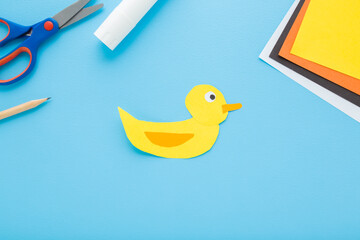 Wall Mural - Created and glued yellow duck shape. Scissors, glue stick, pencil and colorful application paper on light blue table background. Pastel color. Closeup. Child handcraft. Top down view.