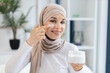 © sofiko14 - Smiling adult Muslim woman wearing hijab applying moisturizer cream on facial skin indoors. Promotes skincare, self-care, beauty regime, and wellness.