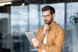 © Liubomir - Businessman standing in a modern office, concentrating on information displayed on a digital tablet, reflecting on strategy, and planning future actions