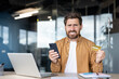 © Liubomir - Young man at office desk holds smartphone and credit card, frowning in frustration over a failed online payment or banking error, stressed and worried about finances