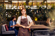 © bnenin - Confident Female Employee in Apron Standing in Stylish Outdoor Cafe Ambiance