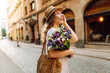 Cheerful woman in hat strolls along sunny street with bouquet of wildflowers. Beautiful woman holds bouquet and enjoys city atmosphere. Conceptual concepts for walking, recreation, and tourism.