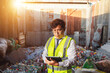 © pichet - Professional waste management worker using tablet device in recycling facility surrounded by plastic bottles and containers in urban environment
