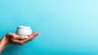 © Dulemegapixel - A hand elegantly holds a jar of luxurious cream against a calming blue background, highlighting skincare and beauty, evoking feelings of relaxation and indulgence.