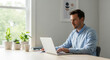 © yahya - A focused man in a light blue shirt works diligently on his laptop at a bright desk with potted plants, illuminated by natural window light.